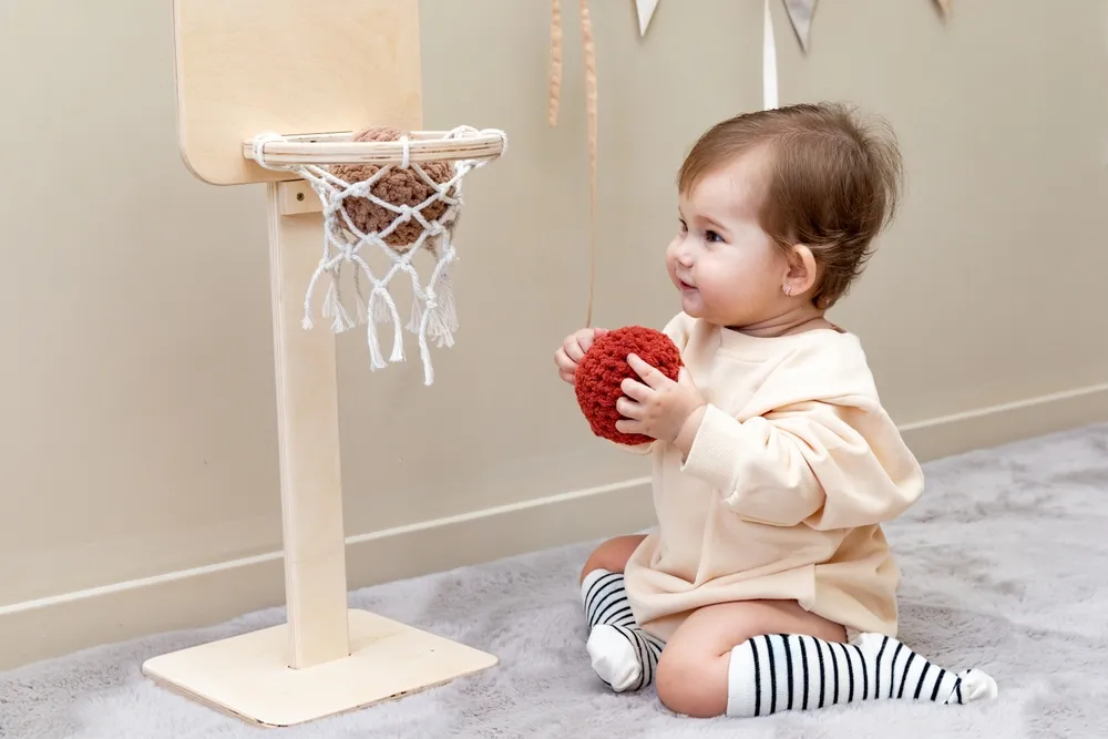 Petite fille en costume beige assise avec un ballon souple sur le sol près d'un panier en bois et jouant au basket-ball dans une chambre